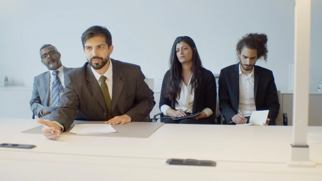 Hiring panel of four seated at a desk conducting a job interview, with one panelist speaking and others taking notes